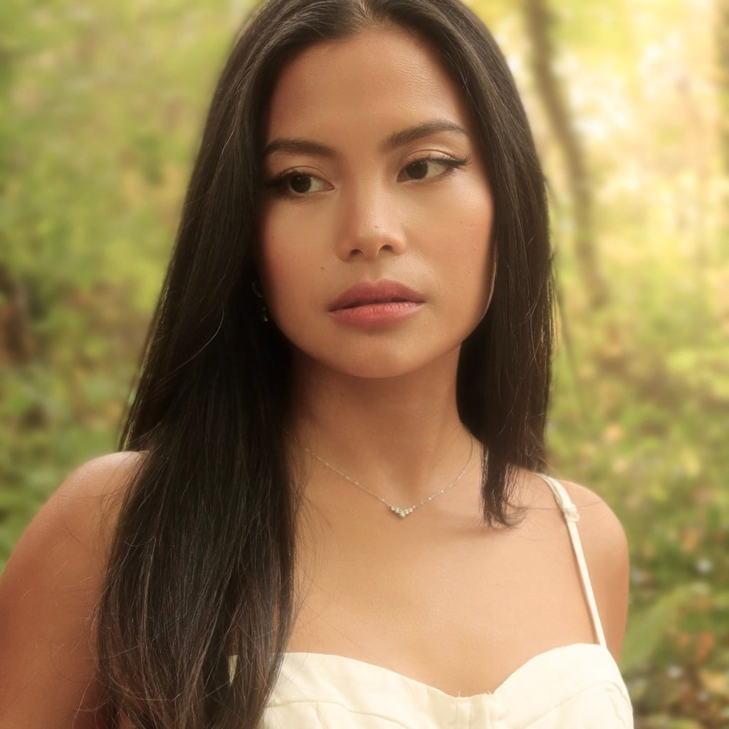 Outdoor portrait of a woman wearing a sterling silver V-shaped cubic zirconia necklace, standing in soft golden forest light. Her long dark hair and ivory dress frame the delicate pendant, which catches the natural light against the blurred woodland background.