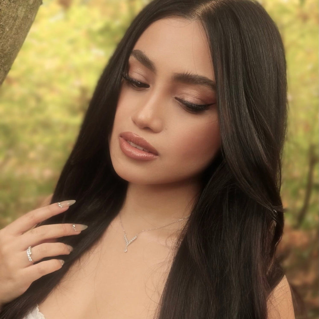 Soft outdoor portrait of a woman wearing a sterling silver Y-shaped cubic zirconia necklace and matching ring, standing in warm forest light. Her long dark hair and white dress frame the delicate pendant, which sparkles subtly against the blurred woodland background.