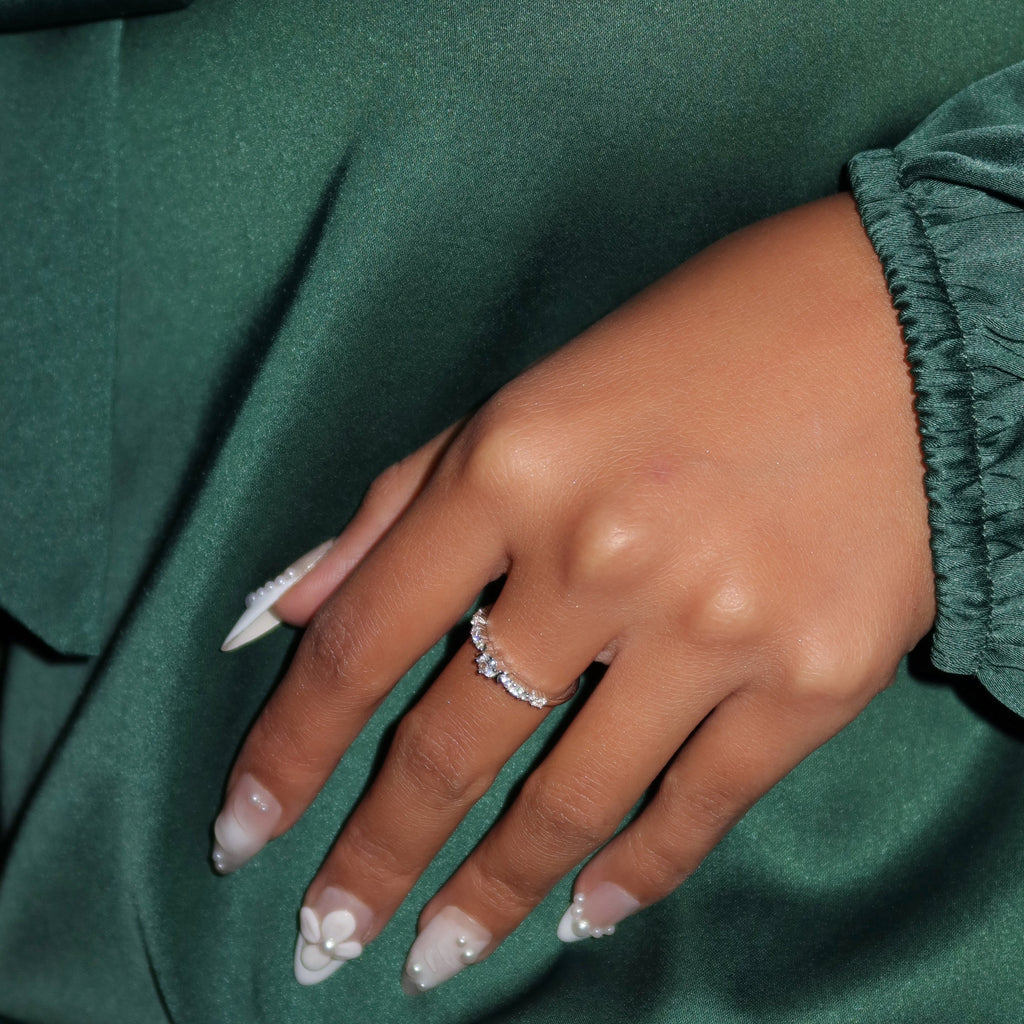 Close-up of a woman wearing a sterling silver eternity ring with sparkling round stones, her hand resting on deep green satin fabric while showcasing pearl-accented floral nails.