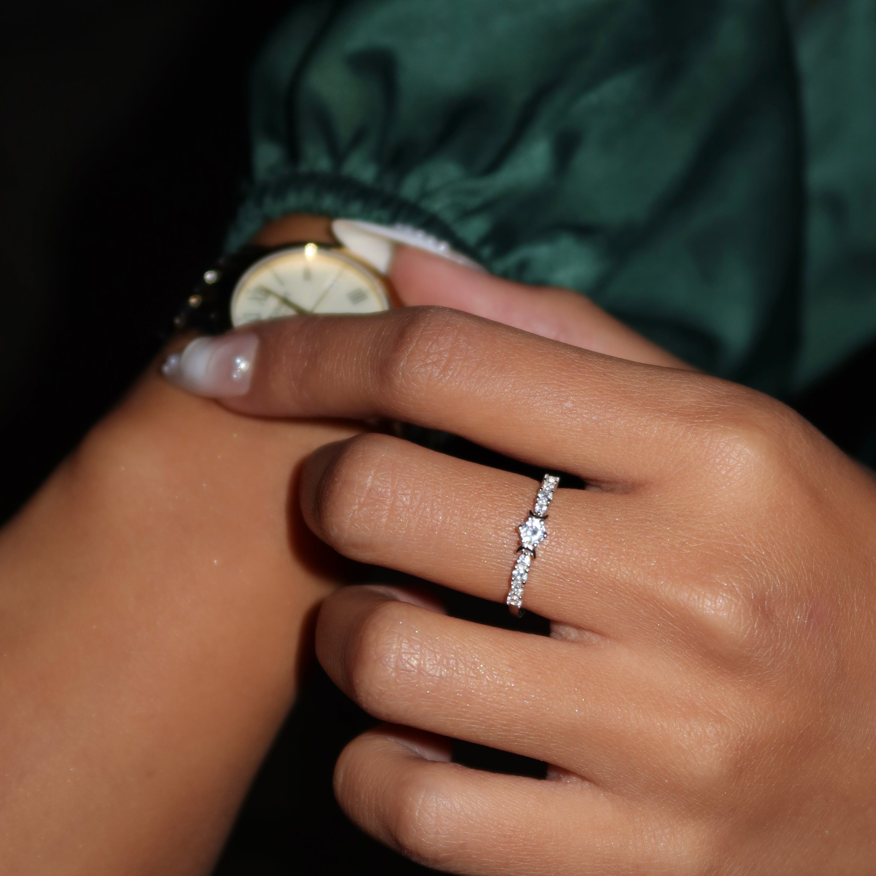 Close-up of a woman wearing a delicate sterling silver ring with a small round center stone and pavé detailing, her hand adjusting a gold wristwatch while dressed in a green satin sleeve.