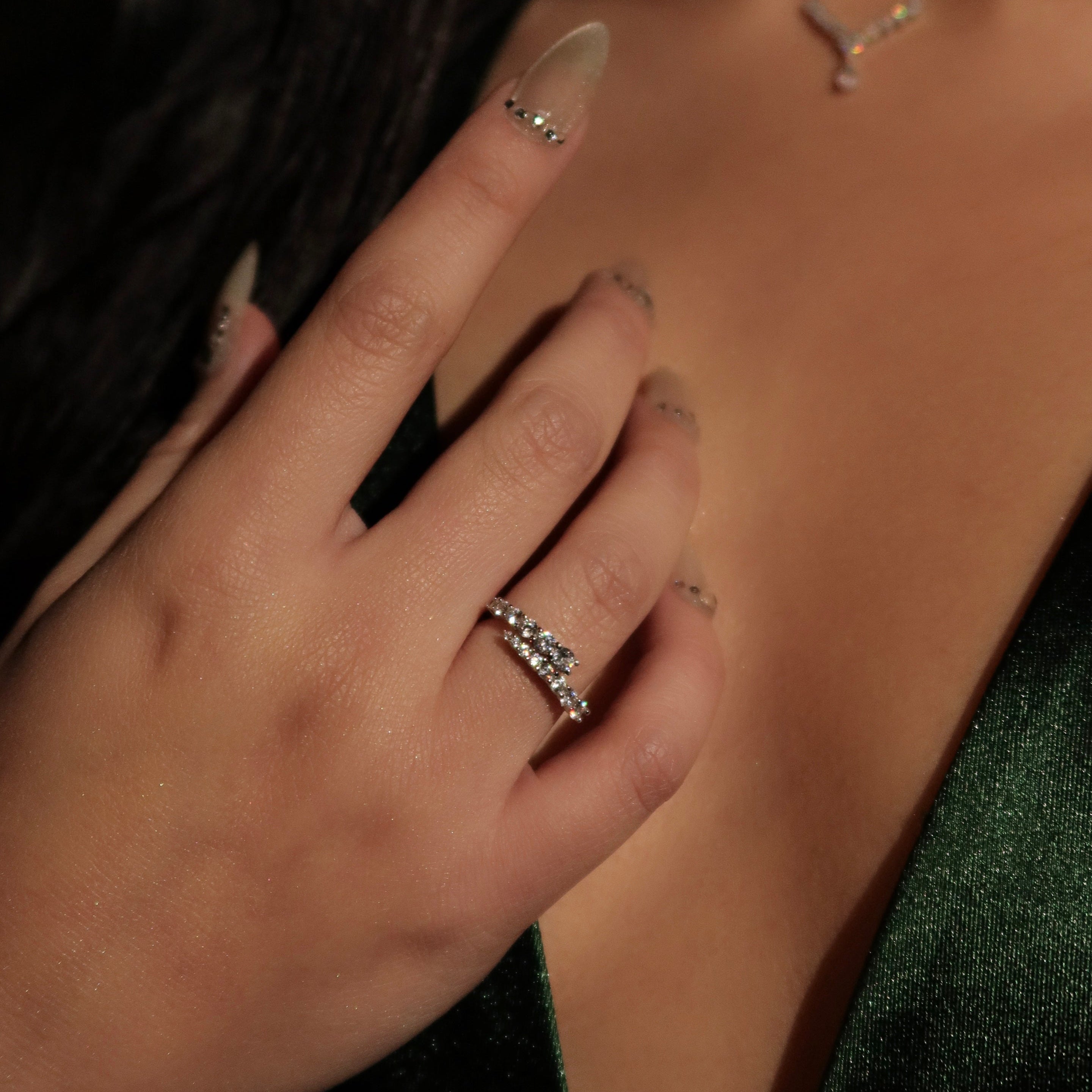 Close-up of a woman wearing stacked sterling silver rings with sparkling round stones, her hand resting near her neckline while dressed in a deep green satin outfit and matching jewelry.
