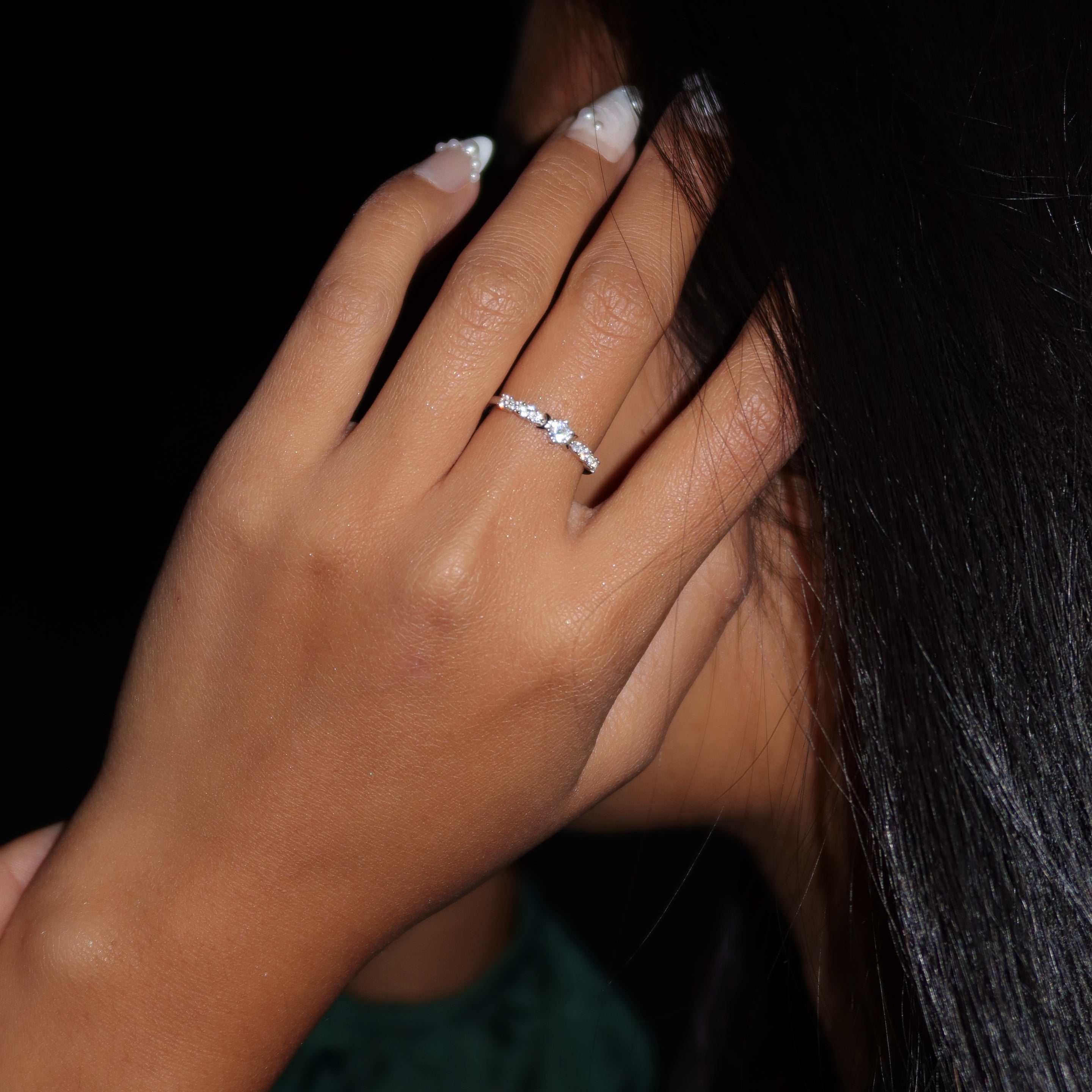 Close-up of a woman wearing a delicate sterling silver ring with a small round center stone and pavé band, her hand gently brushing her hair in a dark, softly lit setting.