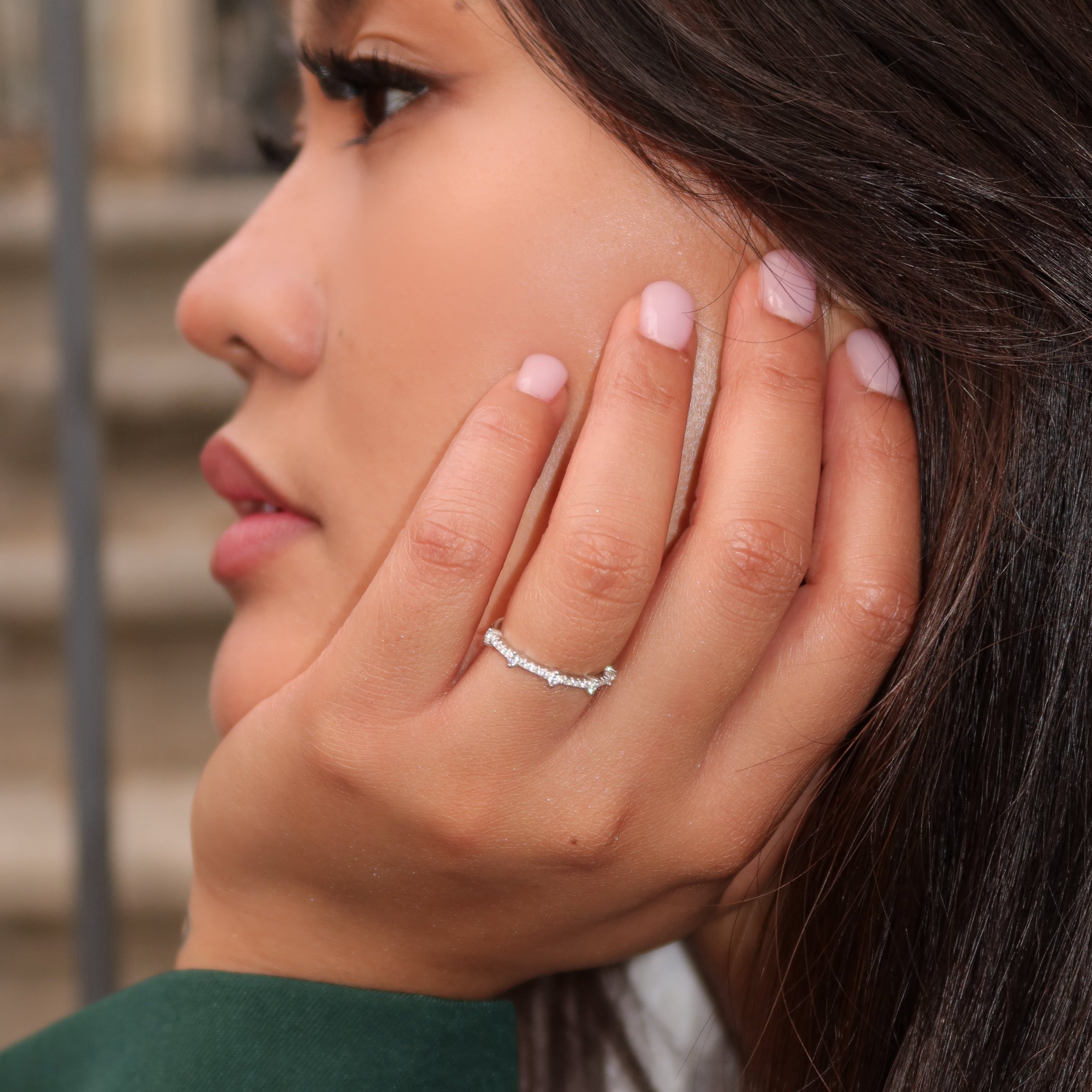 Close-up of a woman wearing a delicate sterling silver band with small round stones, her hand resting against her face with soft pink nails and long dark hair.