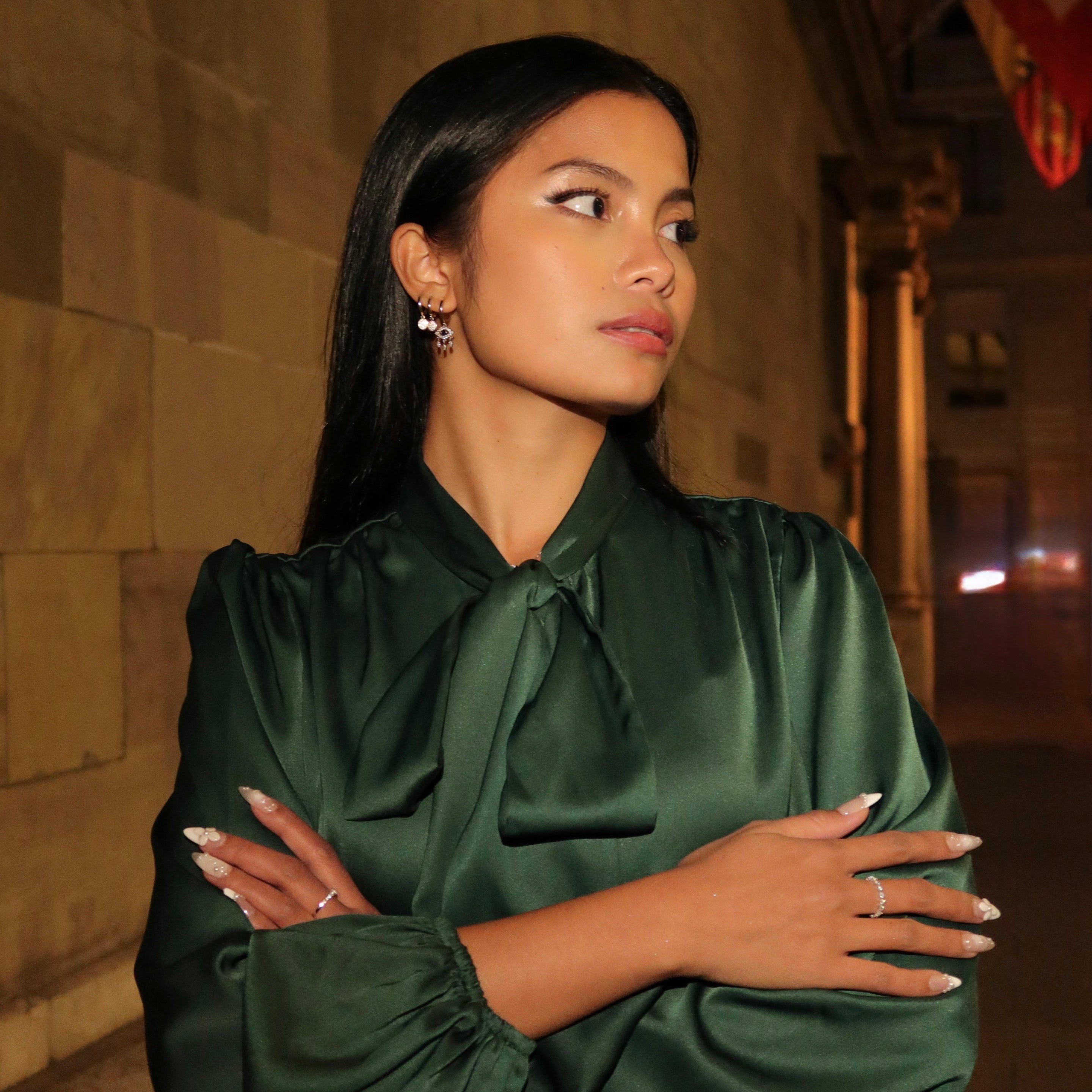 Woman wearing sterling silver earrings and a delicate eternity ring, posing in a green satin blouse on a dimly lit street with Swiss flags in the background.