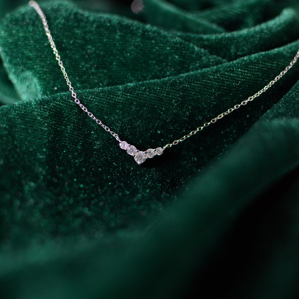 Close-up photograph of a sterling silver V-shaped necklace resting on deep green velvet. The delicate chain and clustered cubic zirconia stones catch the light softly, creating an elegant sparkle against the rich velvet backdrop.