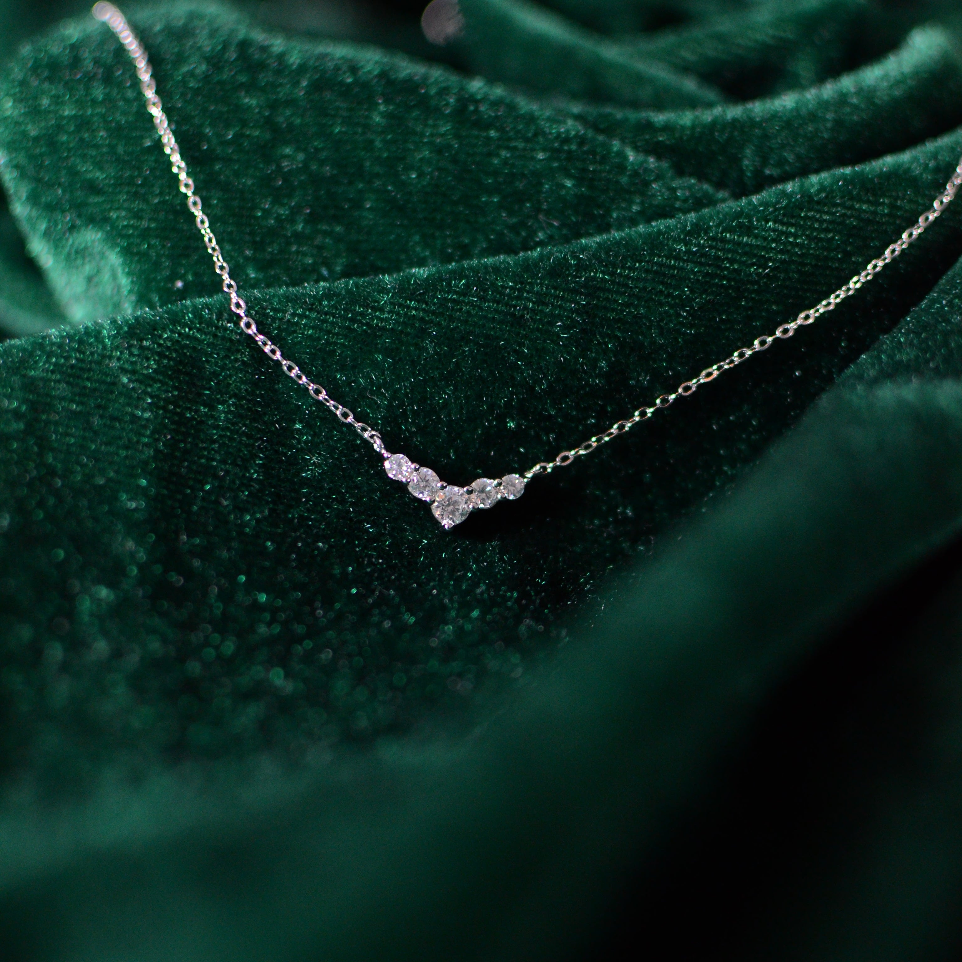 Close-up photograph of a sterling silver V-shaped necklace resting on deep green velvet. The delicate chain and clustered cubic zirconia stones catch the light softly, creating an elegant sparkle against the rich velvet backdrop.