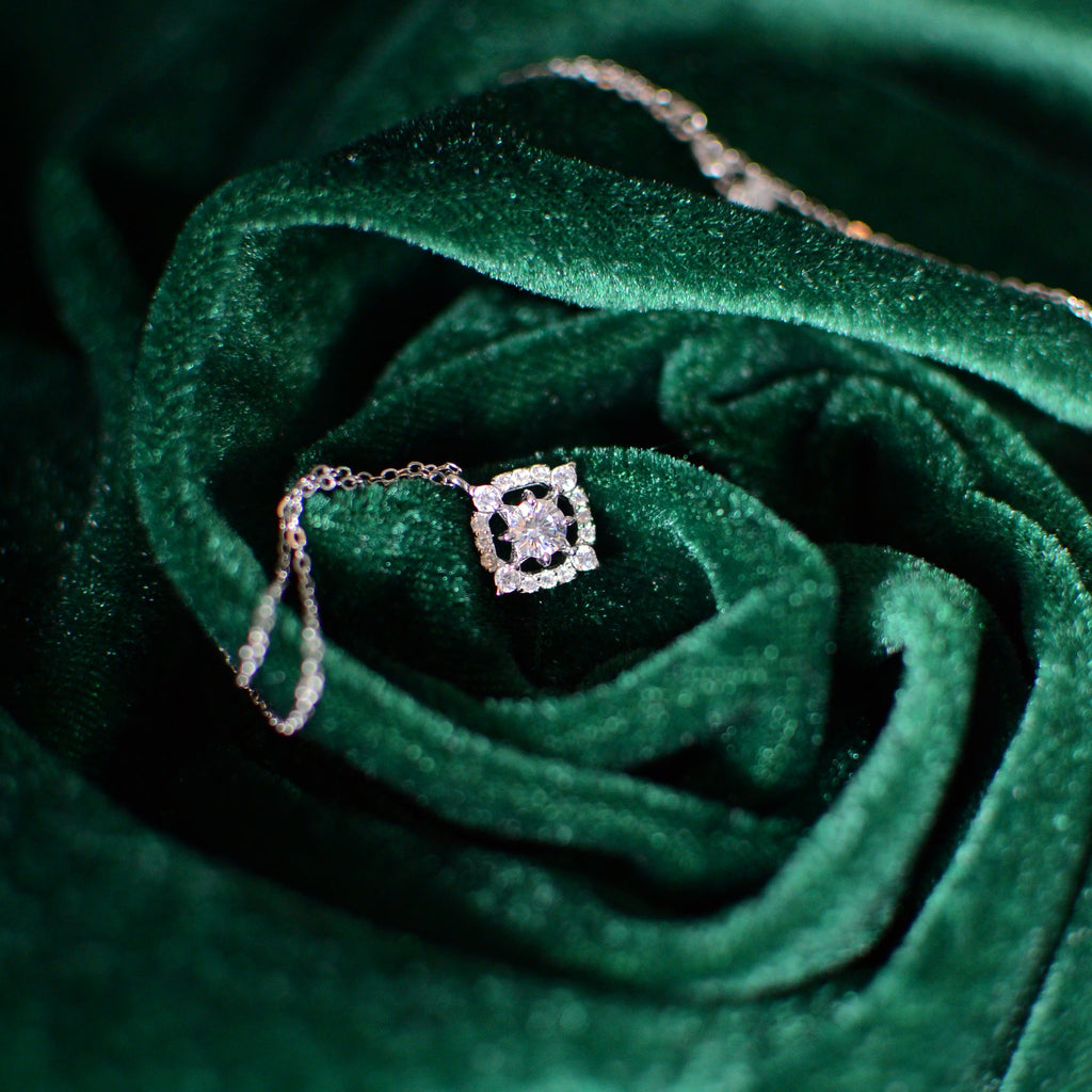 A square-halo sterling-silver pendant necklace with sparkling cubic zirconia stones resting on deep green velvet folds, highlighting the pendant’s brilliance and fine chain in a rich, luxurious setting.