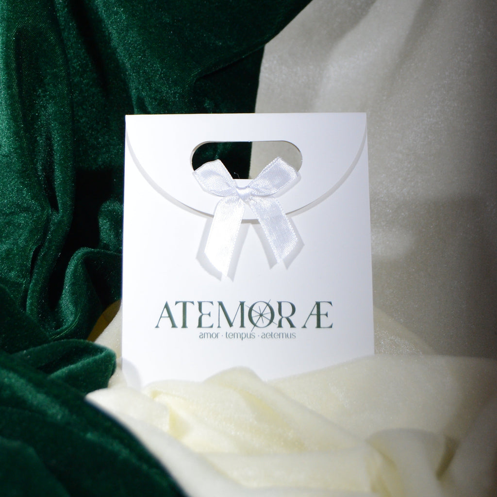 Atemorae white gift bag with a satin ribbon bow, displayed against deep green and cream velvet fabric for a refined, elegant presentation.