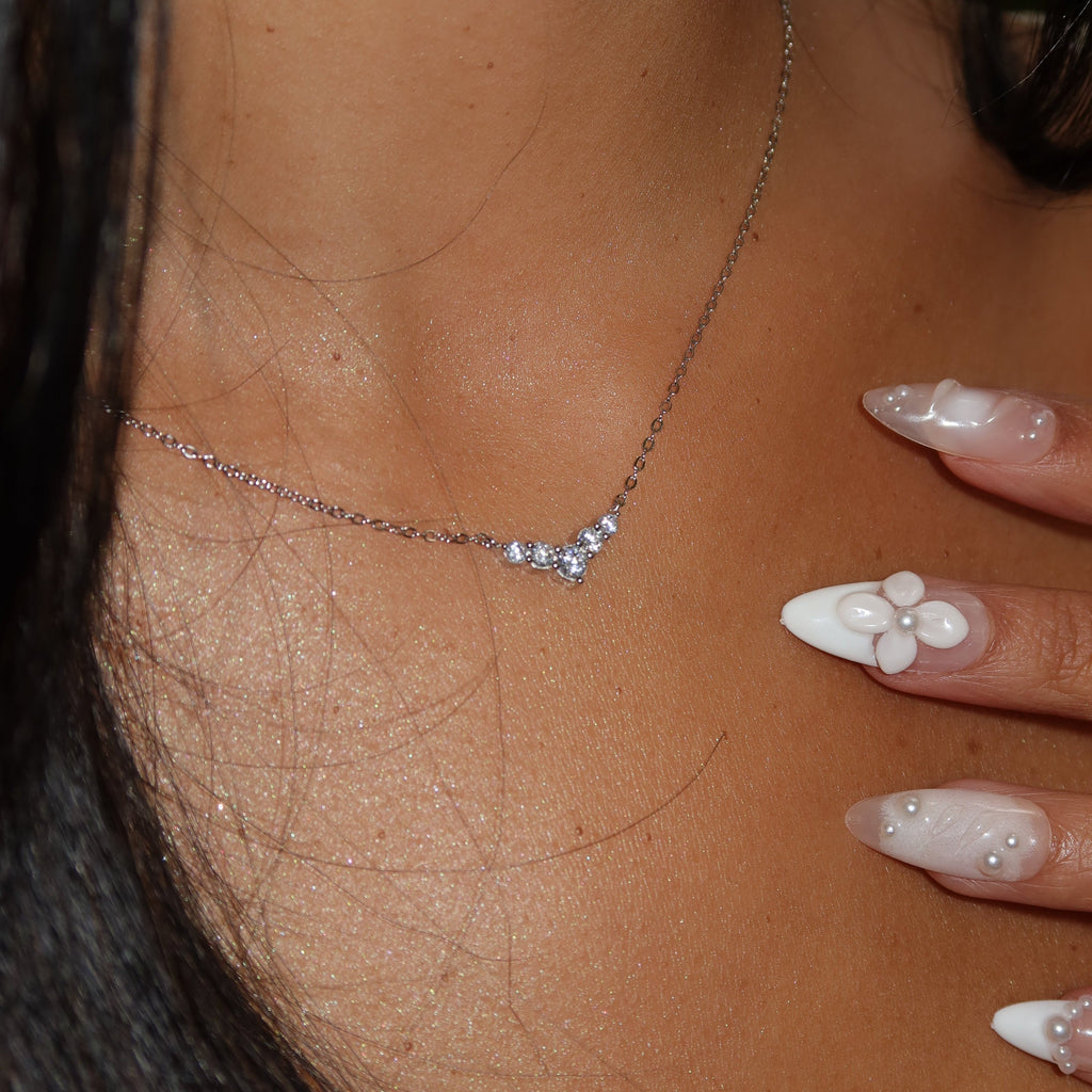 Close-up view of a sterling silver V-shaped cubic zirconia necklace resting on a woman’s collarbone, highlighted by soft shimmer on the skin and pearl-accented white nails touching the pendant for emphasis.