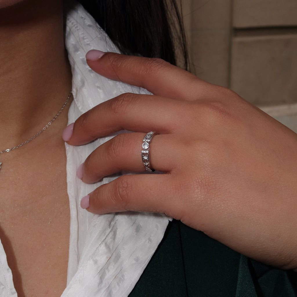 Close-up of a woman wearing a sterling silver ring with a round center stone and detailed side accents, her hand resting on her chest while dressed in a white textured top and dark green jacket.