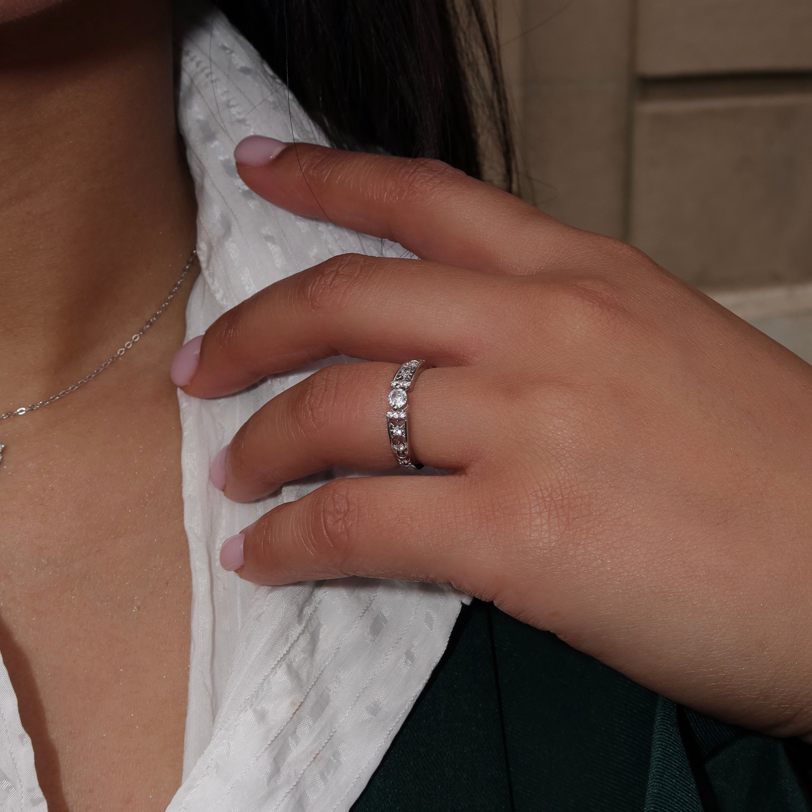 Close-up of a woman wearing a sterling silver ring with a round center stone and detailed side accents, her hand resting on her chest while dressed in a white textured top and dark green jacket.