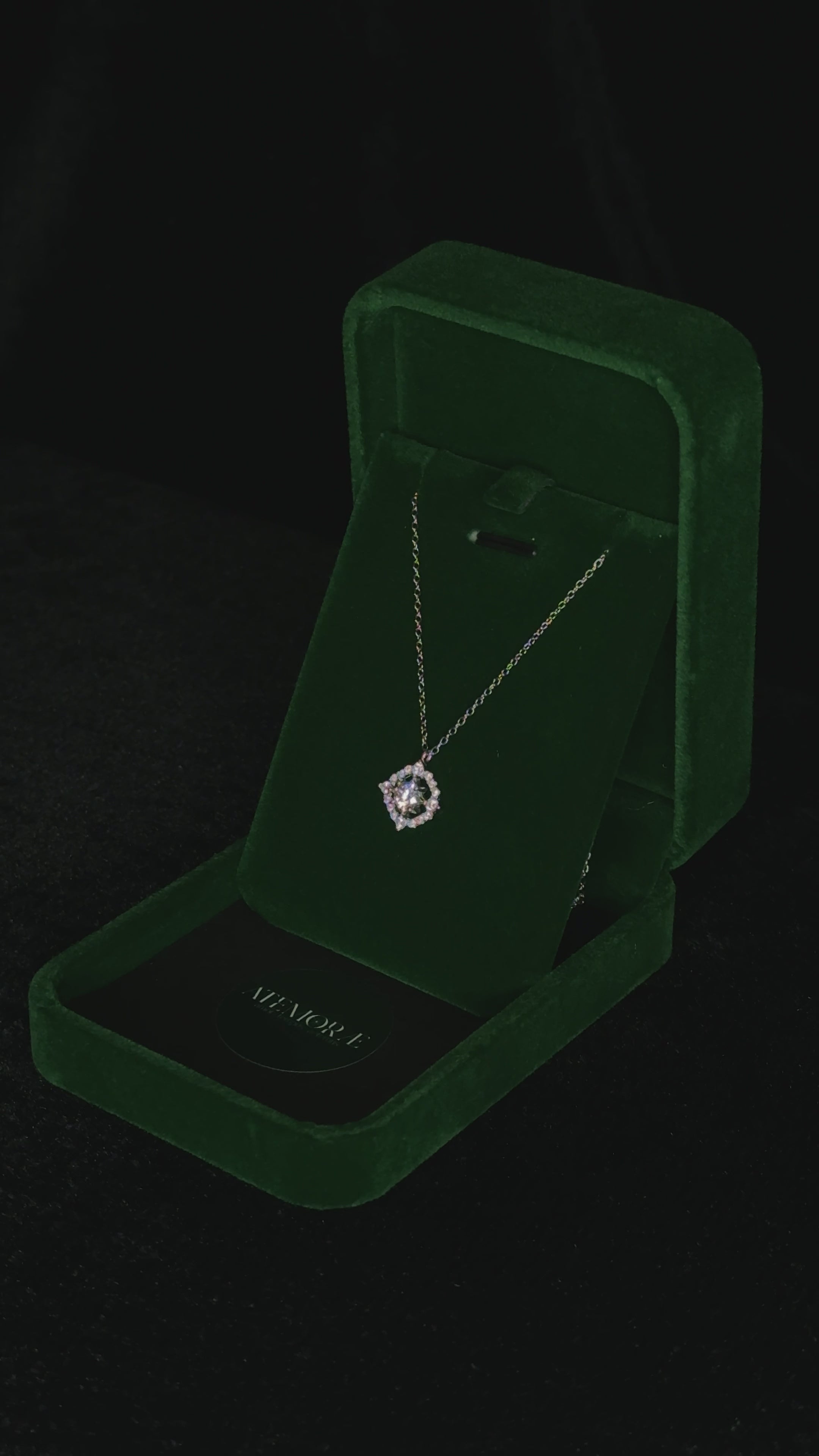 Close-up video of a sterling silver halo pendant necklace gently rotating inside a deep green velvet jewelry box. The square halo design catches the light from every angle, revealing the brilliant sparkle of the cubic zirconia stones and the fine detailing of the chain.